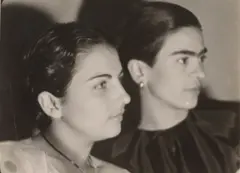 Perfil do rosto de Cristina, que usa uma blusa branca e um colar. Ao seu lado, ao fundo, está Frida, que usa uma blusa preta e brincos. Ela também aparece de perfil