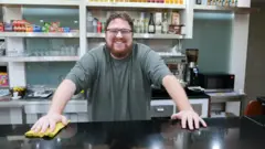 Daniel Silva, de óculos e barba e cabelos ruivos, sorrindo atrás de um balcão preto, cobre o qual ele segura um pano amarelo. Atrás dele se vê uma máquina de café expresso, copos e taças de vidro, e alguns alimentos