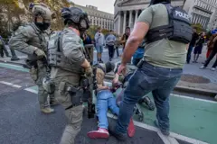 Agentes de polícia fortemente armados detêm uma pessoa no chão durante operação em rua urbana, com pessoas observando ao fundo