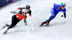 Buz pateni yapan üç erkek sporcu, soldaki ay yıldızlı, sağdaki mavi ve italyan bayraklı bir üniforma giyinmiş, ortadaki sporcu soldakinin arkasında sadece kaskı yüzü ve kolları görünüyor, üçü de zor bir virajdan hızla geçerken yan yatmış görünüyorlar. 