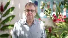 Homem branco, de óculos, com camisa cinza e flores roxas à direita e ao fundo