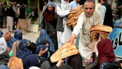 یک مرد افغان با لباس سفید در مقابل یک نانوایی در حال توزیع نان برای شماری از زنان دیده می شود