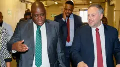 Home Affairs Minister Leon Schreiber wear white tie and di Deputy Nzuza wear green tie wit suit wen dem visit di govment Printing Works