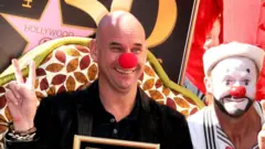 Guy Laliberté attends the Ceremony honouring him with a star on the Hollywood Walk of Fame on 22 November. He is wearing a red clown’s nose and has his hands in a V- sign. To his left is a male clown with a white face paint, red nose, and a black beard and moustache painted into his face. The Hollywood Star is on a plaque behind them and Laliberté is holding a picture frame with the star inside.