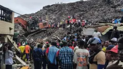 Rescuers use an excavator to comb through a mountain of scattered rubbish at a major waste site in Conakry, Guinea, as local residents gather nearby. Photo: 23 August 2026