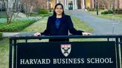 تقف شريا ميشرا ريدي داخل حرم هارفارد الجامعي في ولاية بوسطن في الولايات المتحدة، خلف لافتة تحمل عبارة "كلية هارفارد للأعمال".