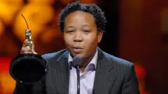 Presley Chweneyagae dey deliva a speech afta e win di Oscar Outstanding Performance by an Actor for a Leading Role for "Tsotsi". E dey lift im award up as e wear black stripe suit