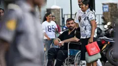 Tekerlekli sandalyede sakallı bir kişi itiliyor. Fotoğrafta başka kişiler de var.