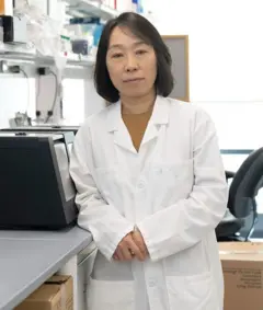 A professora Xu Yu veste um jaleco branco e olha para a câmera com um olhar sereno, ao lado de um equipamento de laboratório