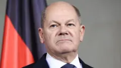 Foto do chanceler alemão Olaf Scholz, inexpressivo1xbet quem é o donofrente a uma bandeira vermelha e preta