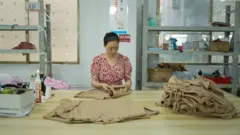 Uma mulher de vestido vermelho está em pé em uma grande mesa de madeira dobrando camisetas marrons. Um óleo de calças marrons dobradas também está sobre a mesa.