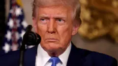 O presidente dos EUA, Donald Trump, um homem branco idoso com bronzeamento artificial e cabelos loiros, vestindo um terno azul-marinho com gravada azul com o semblante sisudo