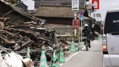 車道沿いの家屋が地震で倒壊しており、緑色のコーンで封鎖されている。その横を車や自転車が走っている