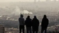 4 رجال يظهرون من الخلف حيث يراقبون من سفح تل،  عموداً من الدخان يتصاعد عقب انفجار وقع في 2 مارس/آذار 2026 في طهران، إيران.