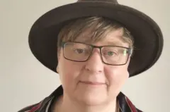 Usando um chapéu marrom de aba larga e óculos pretos, a cineasta Rachel Dax olha em direção à câmera.