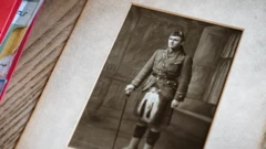 Fotografia em papel, em preto e branco, de James Grant Allan, de pé com kilt e sporran escocês e boné militar. A fotografia está emoldurada e se encontra sobre uma mesa de madeira, com uma pasta vermelha à esquerda.