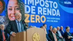 Lula sorri ao discursar em um púlpito da presidência da República, com integrantes do governo sentados atrás. Atrás de Lula, há também um banner com uma mulher sorridente e a mensagem: 'IMPOSTO DE RENDA ZERO'.