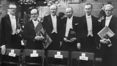 Vestidos de gala, vencedores do Prêmio Nobel de 1962 exibem seus diplomas após as cerimônias na Sala de Concertos de Estocolmo, na Suécia. Da esquerda para a direita, Maurice H. Wilkins (Medicina, 1916-2004); Max Perutz (Química, 1914-2002); Francis Crick (Medicina, 1916-2004); John Steinbeck (Literatura, 1902-1968); James D. Watson (Medicina, 1928-2025); e John Kendrew (Química, 1917-1997).