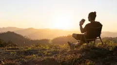 muškarac sedi na stolici na uzvišenju okrenut kameri, u ruci drži kafu dok gleda u sunce koje izlazi