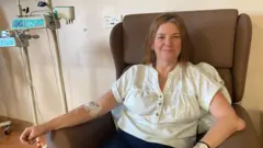 Une femme vêtue d'une chemise blanche est assise dans un fauteuil marron. Elle reçoit une perfusion de plasma par le biais d'un cathéter inséré dans son bras. Des pansements recouvrent le point d'entrée du cathéter à l'intérieur de son coude. À sa gauche se tient un technicien hospitalier.