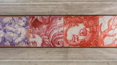 Um painel horizontal reúne desenhos em tons de vermelho e azul que remetem a figuras humanas, rostos e formas orgânicas em movimento. A obra, instalada sob uma estrutura de concreto aparente, mistura referências aos azulejos tradicionais com traços fluidos e fragmentados.