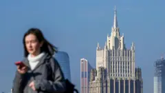 На заднем плане многоэтажное здание со шпилем на фоне голубого неба. На переднем плане в левой части размытая фотография девушки по пояс. Девушка идет справа налево. У нее темные длинные волосы. Она одета в серую куртку и белый свитер. На левом плече у девушки черная сумка. В правой руке - красный смартфон.