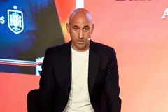 Luis Rubiales olha para a frente, com a cabeça inclinada. Ele está sentado e usa um terno preto e uma blusa branca