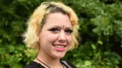 Kayla, com cabelos loiros curtos e piercings no rosto, sorrindo para a câmera