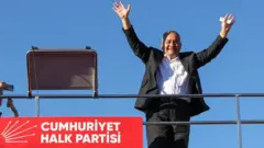 22 Ağustos 2025. Özarslan siyah takım elbiseli ve beyaz gömlekli bir otobüsün üzerinden ellerini kaldırmış selam veriyor. Sol altta CHP logosu görünüyor. 