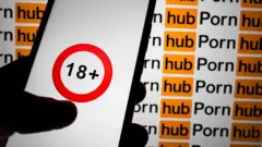 Dłoń trzymająca telefon komórkowy z logo 18+ w czerwonym okręgu. W tle widoczne jest logo strony Pornhub.