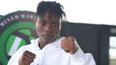La combattante de MMA Juliet Ukah porte un haut blanc et brandit deux poings en regardant droit dans les yeux.