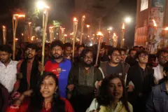 Estudantes, homens e mulheres, caminham de braços dados com tochas acesas, nas proximidades da Universidade de Daca, no domingo (16/11/2025)
