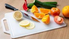 Frutas e legumes diversos, alguns deles cortados com uma faca, sobre uma tábua de corte de plástico