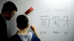 Aluno copia os ideogramas da palavra "Portugal" em mandarim em um quadro branco, sob a supervisão do professor