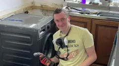 Um homem está sentado ao lado de uma máquina de lavar, segurando uma furadeira enquanto realiza reparos. Ele veste uma camisa da banda Sex Pistols.