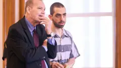 Хади Матар в полосатой робе и наручниках говорит со своим адвокатом в зале суда