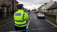 Um policial usando um colete de alta visibilidade atrás de uma fita de isolamento azul e branca que bloqueia uma rua residencial em Glasgow