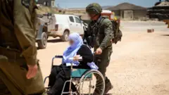 رجل يرتدي الزي العسكري الإسرائيلي يحرك كرسياً تجلس عليه سيدة مسنة.
