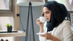 Une femme voilée boit du café dans une petite tasse blanche, son voile est noir, sa chemise est blanche et elle porte une veste de soirée blanche par-dessus.