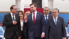 政府専用機を降りたスーツ姿のヴァンス氏が、スーツ姿のパキスタン高官たちに囲まれ、右手の人差し指を立てて話しながら前へ向かって歩いている