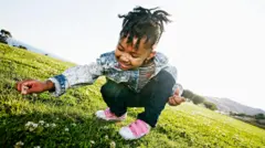 Una niña sonríe mientras está agachada en cuclillas profundas sobre un campo de césped iluminado por el sol. Está buscando pequeñas flores blancas. La niña lleva una chaqueta vaquera, pantalones oscuros y zapatos rosas.