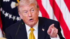 トランプ氏がマイクの前で、左手の人差し指で何かを指さしながら話している。濃い色のスーツに白いシャツ、黄色のネクタイを着用し、襟には星条旗のバッジを付けている