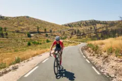 Uma pessoa em uma bicicleta, em um ensolarado dia de verão, desce uma estrada sinuosa que vem de dois altos montes.