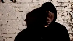 Мужчина в черной одежде с каплюшоном, лица почти не видно, т.к. оно закрыто платком. Он стоит у стены из светлого кирпича.