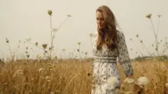 كيت ترتدي فستاناً أبيض مزركش باللون الأزرق في حقل من القمح والورد، الصورة مأخوذة نهاراً