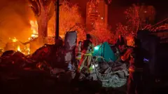 An emergency responder stands amid debris from a strike in Ukraine, lit up by a fire in the distance.