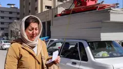 A woman appears to be crying as she walks on a street in Tehran. She has a light coloured headscarf and a tan coat on. She is holding a piece of paper in her hand and has a rucksack on her shoulder. A motorcyclist is behind her and cars line the street to her left.