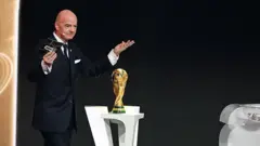Fifa president gesture in front of di trophy. E hold card for hand and wireless mike dey im ear.