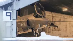 Vista frontal de um caça F-35 dentro de um hangar. Há neve no chão e parece haver também no teto do hangar.