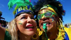 Uma foto em close-up de dois torcedores brasileiros vestindo roupas verdes e amarelas, além de cocares de penas.